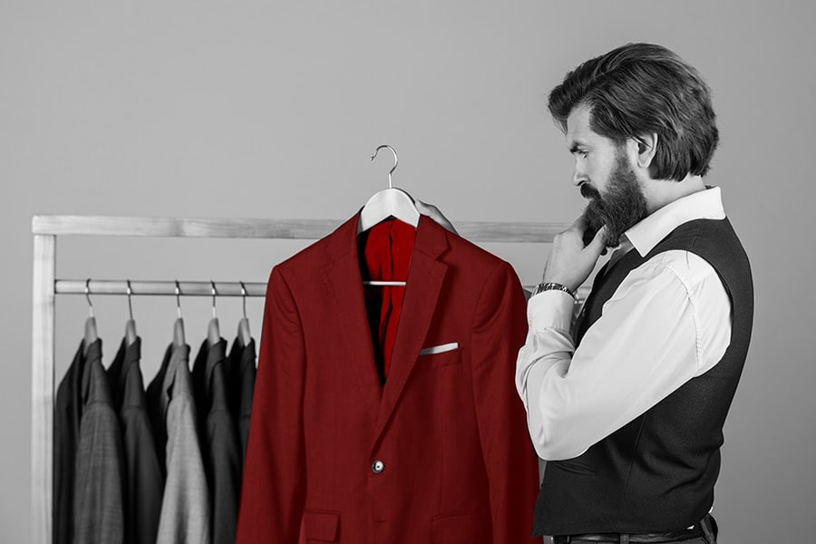 Мъж избира между класически и екстравагантен стил, гледайки червено сако сред редица сиви и черни костюми – визуална метафора за смелия избор и емоционалната сила на моден PR